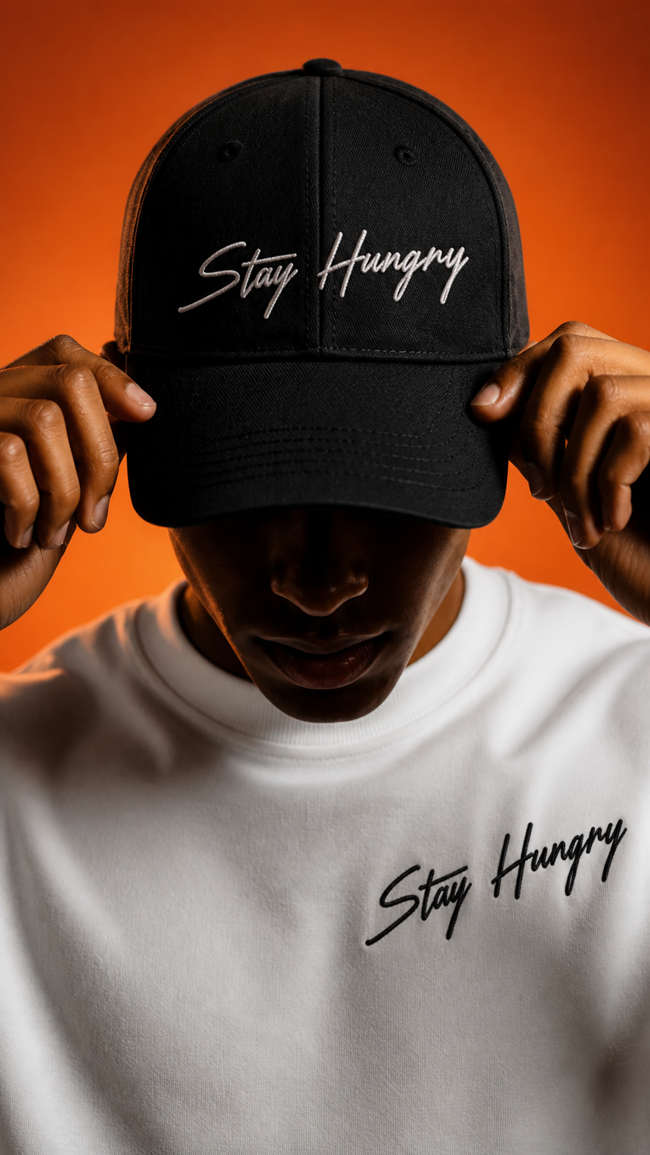 Man pulling down a black Stay Hungry cap against an orange backdrop
